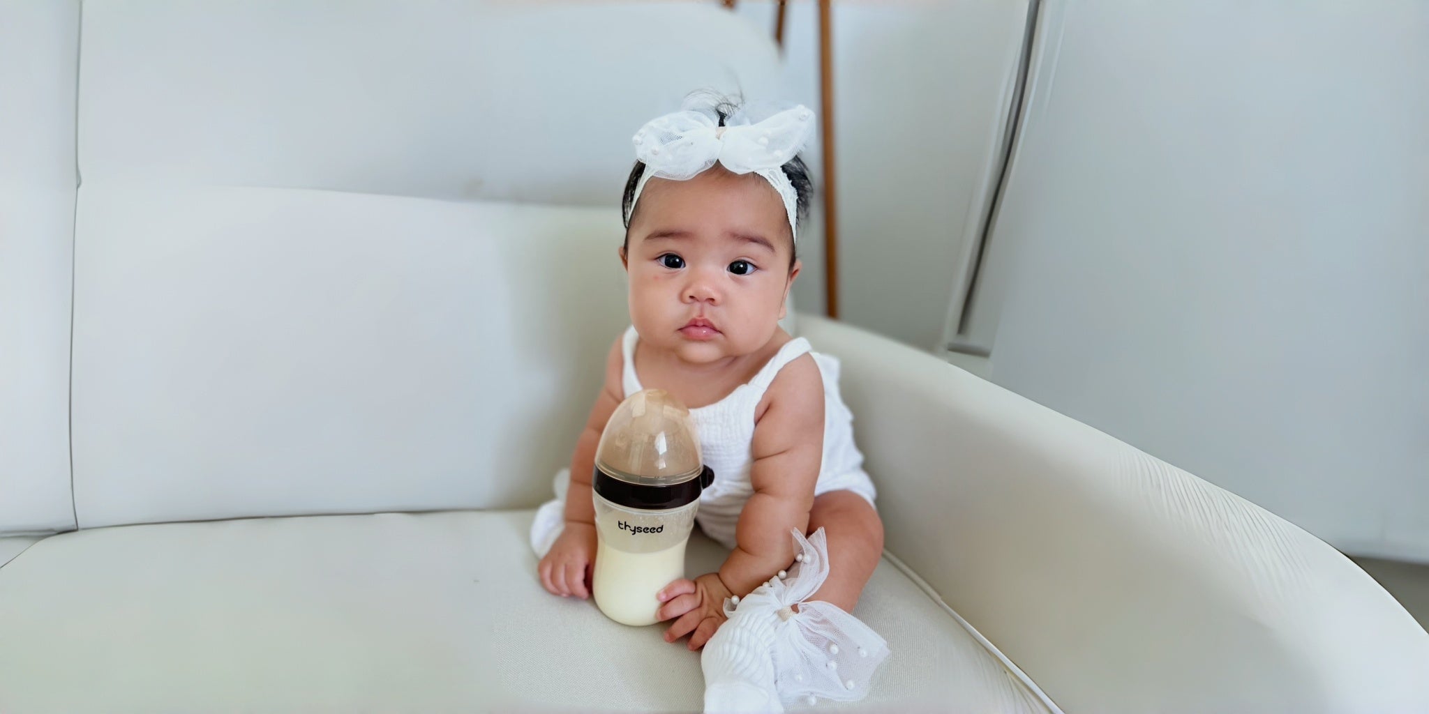 A baby girl sits on a white couch next to a thyseed baby bottle filled with milk.